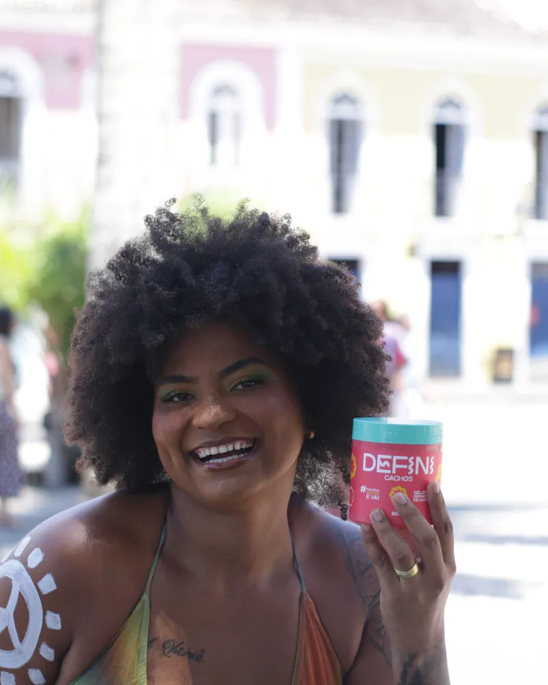 Mulher de cabelo crespo volumoso sorrindo com o pote da Defini Cachos.