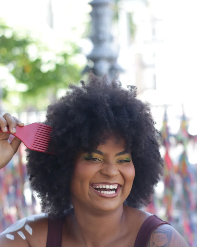 Mulher de cabelo crespo volumoso rindo enquanto ajeita o cabelo com um pente-garfo.