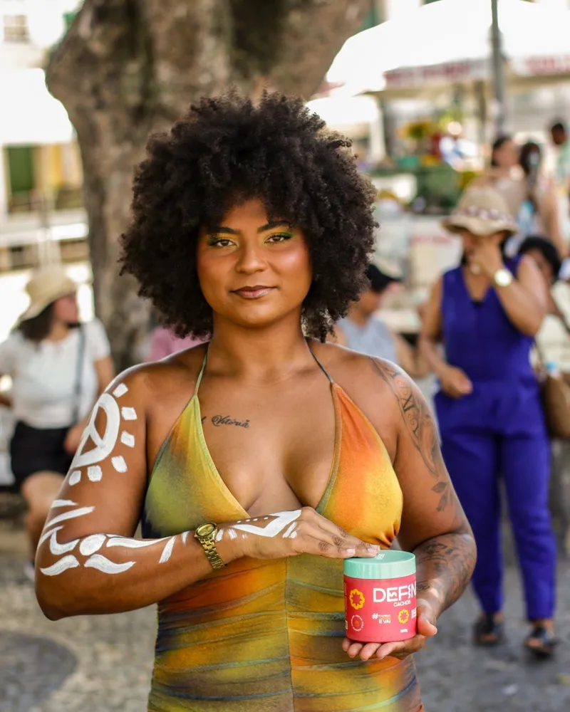 Mulher de cabelo crespo volumoso segurando o pote da Defini Cachos numa praça de Salvador.