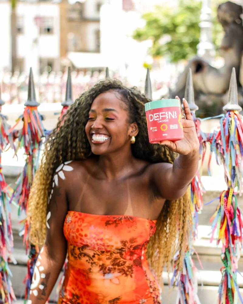 Mulher de cachos longos sorrindo com o pote da Defini Cachos na mão, diante das fitas coloridas em Salvador.