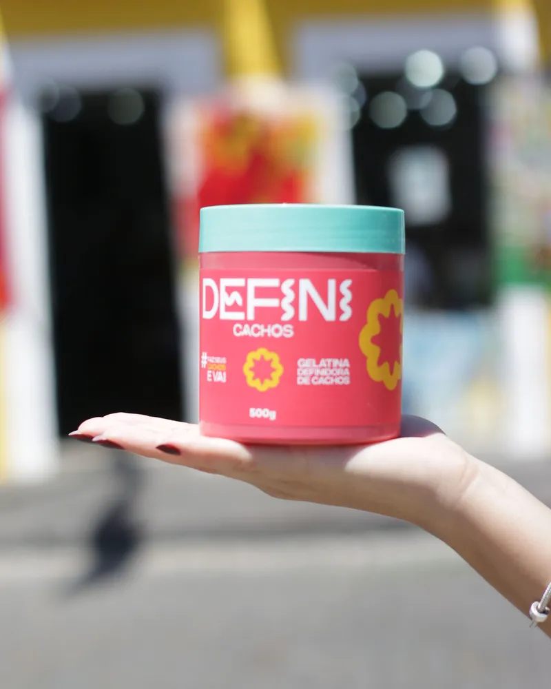 Mão segurando o pote da Defini Cachos, Gelatina Definidora de Cachos 500 g, numa rua de Salvador.